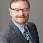 head and chest photo of a man with beard and glasses and short light brownish red hair wearing business attire.