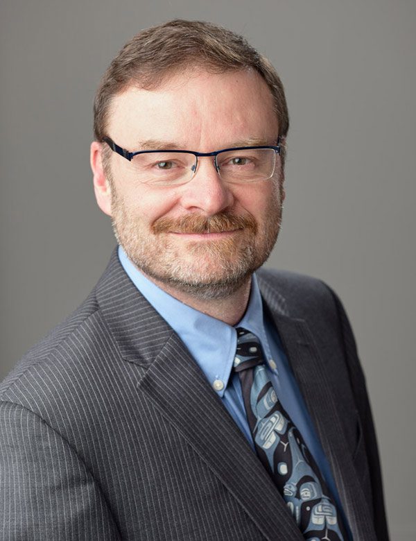 head and chest photo of a man with beard and glasses and short light brownish red hair wearing business attire.