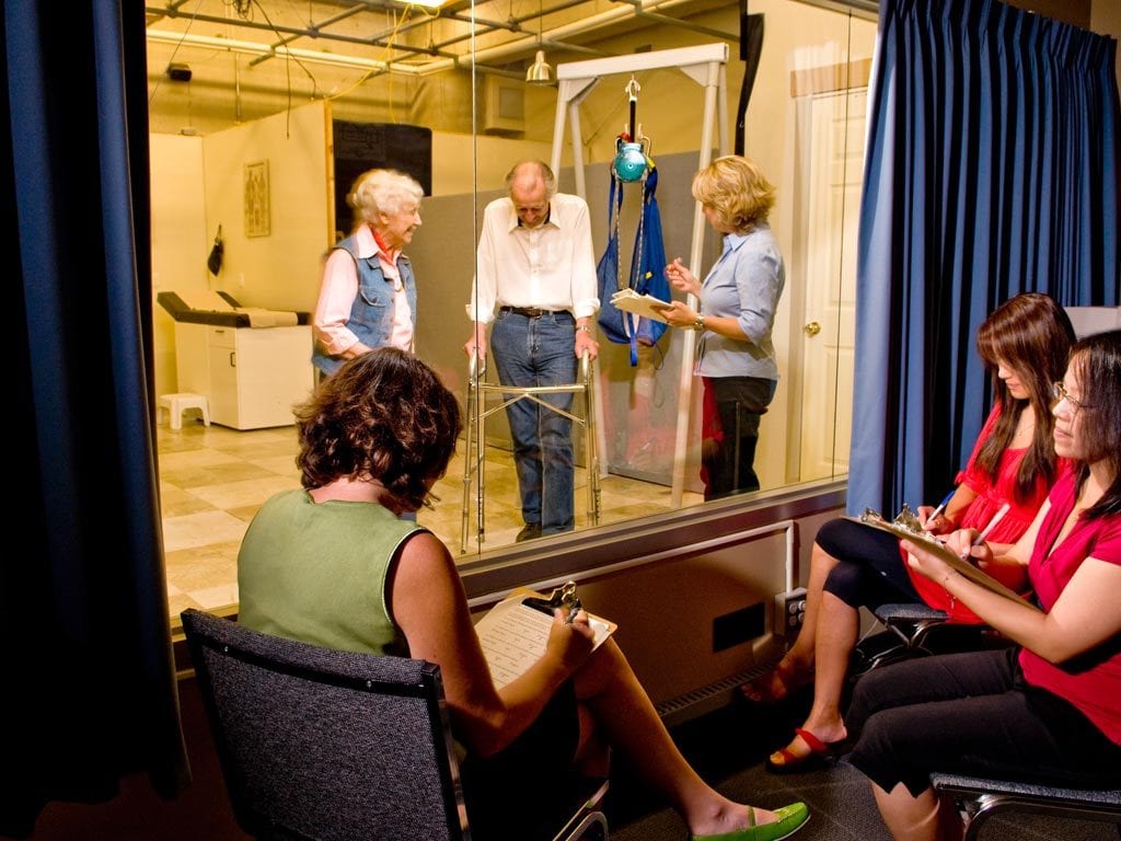 Three female researchers watch three older individuals in a lab from behind a oneway mirror.