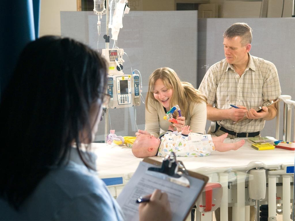 Researcher behind one-sided glass watches woman with baby lying on hospital bed.