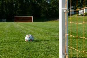 Lawn covered soccer field with two goal nets on each end with a white and orange soccer ball in front of one net.