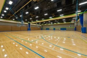 Looking from the entrance there is an empty gymnasium that is well lit with a shiny wood floor with various sport layout markings on the floor.