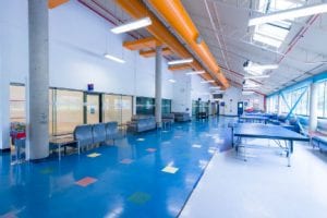 BCIT Student lobby is in a hall-like setting and has a seating area facing squash court windows, ping pong tables along with window area and a blue corridor with overhead yellow air circulation pipes.