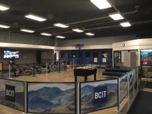 The main lobby of the BCIT Recreation Centre is a classroom size with exercise bikes and equipment along one wall, a foozeball table in centre with seating and is bordered by waist high screens with picturesque mountains and BCIT logo on blue background.