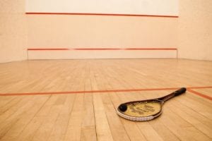 Squash court with beige colored hardwood floor and in the foreground there is a squash racquet with a black ball on the strings.