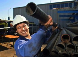 Man with hard hat on lifting a long black pipe off a pile of pipes.