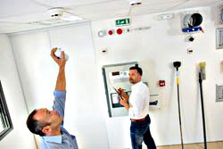 one technician reaching up towards the ceiling in a room and the other technician working at a control panel while looking at the other person.