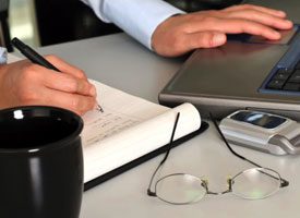 pic of a desking with paper and laptop, cell phone, glasses, coffee mug and a person writing on notepad.