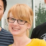 headshot of a young female with black rimmed square glasses, long reddish blond bangs and yellow shirt.