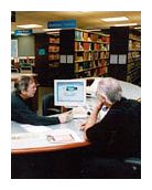 Two people sitting at a table in the library looking at a computer monitor.