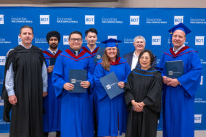 At the 2024 Spring BCIT Convocation ceremony, six TMGT graduates dressed in blue convocation robes stand smiling for a photograph with the BCIT School of Transportation Associate Dean and TMGT BTech Program Head, against a blue and white backdrop printed with the BCIT logo and messaging “Education for a Complex World.” 