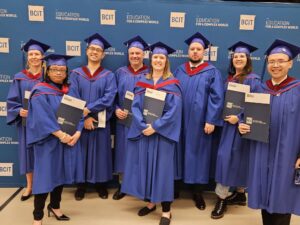 Eight TMGT BTech graduates pose holding their credentials, wearing dark blue convocation gowns and caps in front of a navy backdrop displaying the BCIT logo in white font.