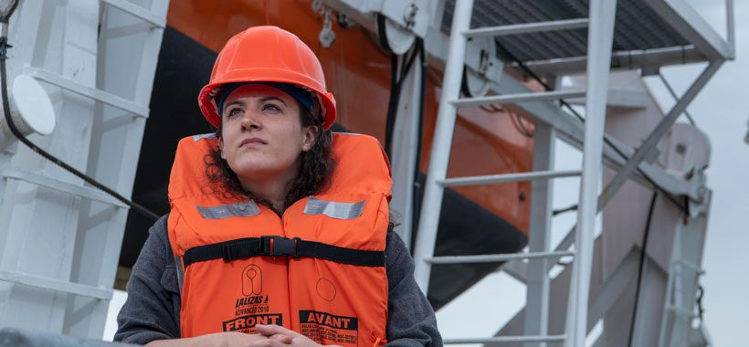 Women dressed as a deckhand in hard hat and life best standing in front of a lifeboat.