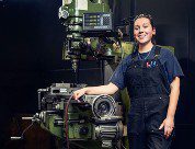 Chelsea Barron - female in blue overalls posing in front of a piece fo machinery 
