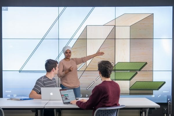 A student presents to two others in front of a large digital screen