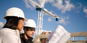 A female and male wearing white hard hats look at blueprints in front of a crane.