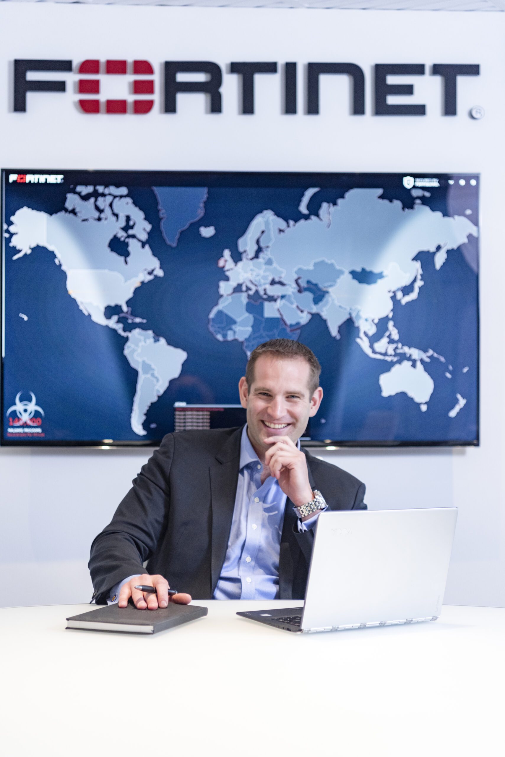 Derek Manky in a boardroom wearing a suit with the Fortinet logo behind him