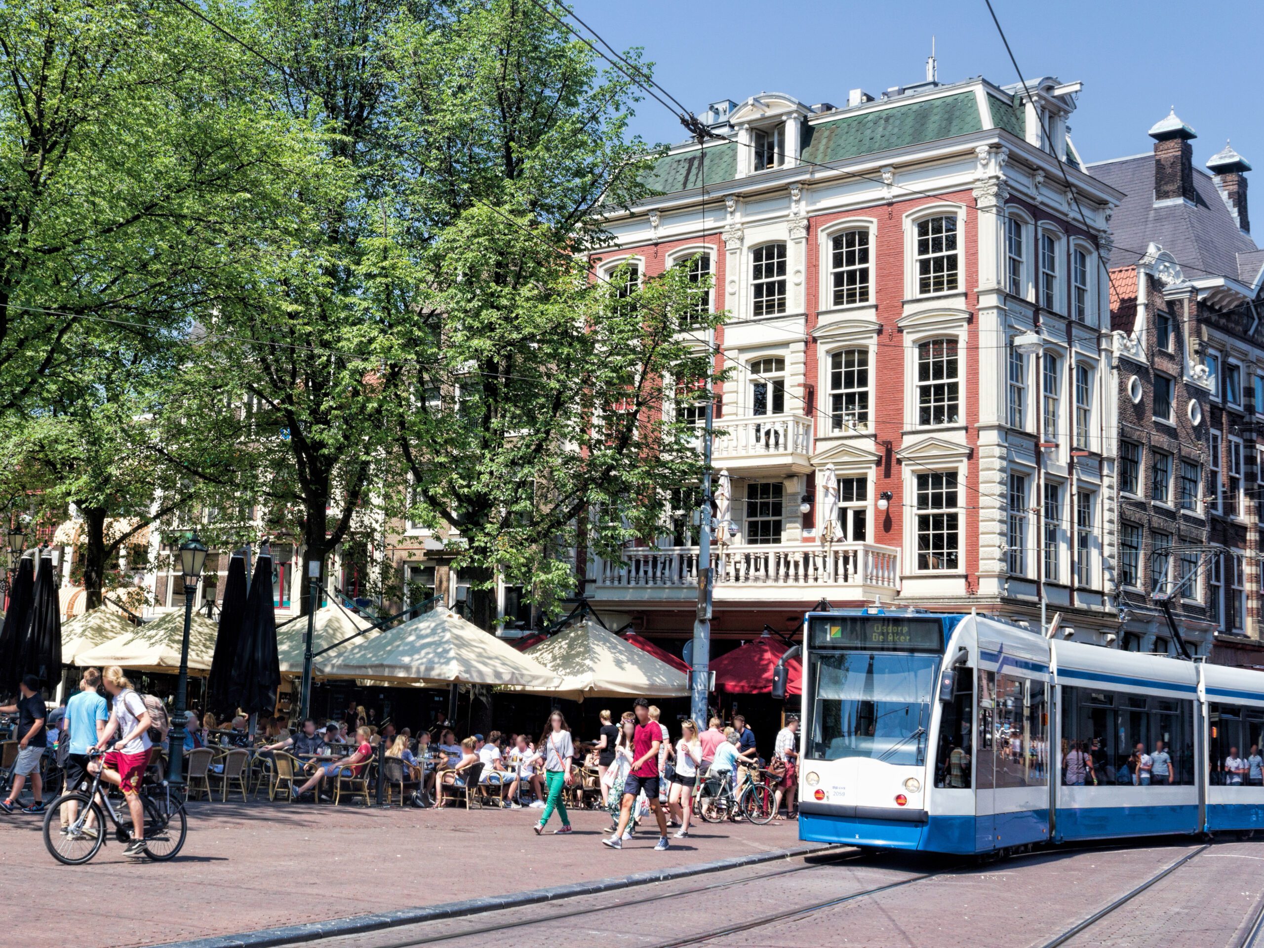 Amsterdam Leidseplein with cyclists, pedistrians, a tram and a full outdoor patio in front of a 5 story brick building.