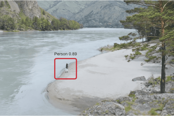 Faraway overhead shot of human figure on the shore of a river with mountains and forest in the background. The figure has a red square around it and reads Person 0.89.