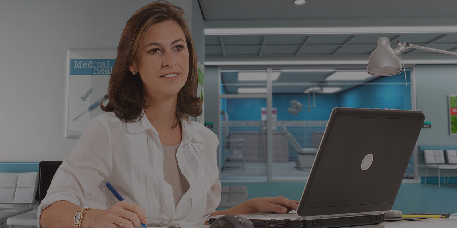 Brunette woman in white shirt working at laptop with medical office equipment behind her