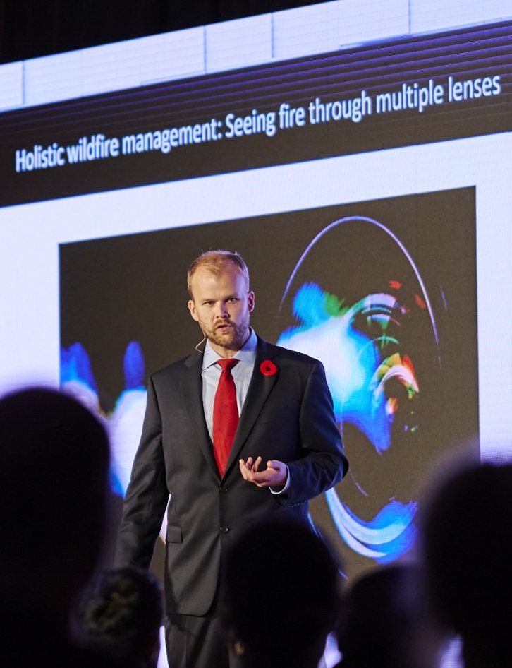 man in suit speaking to an audience with the screen behind him reading "holistic wildfire management: seeing fire through multiple lenses"