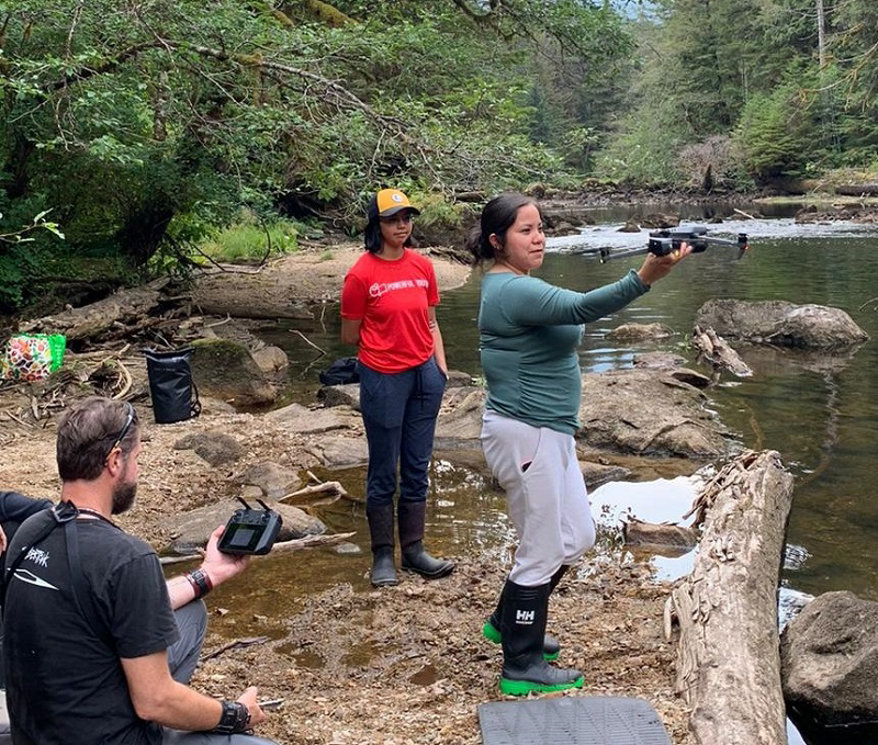 young woman holds out a drone over a forest river, second woman in background and man holding drone controller