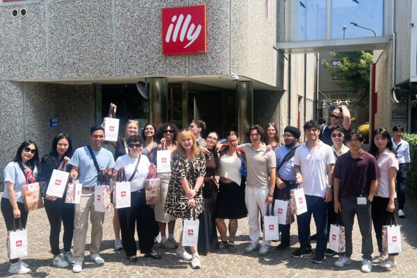 Group of students smiling and standing together in front of an Illy Café company sign, capturing a moment during their study abroad experience